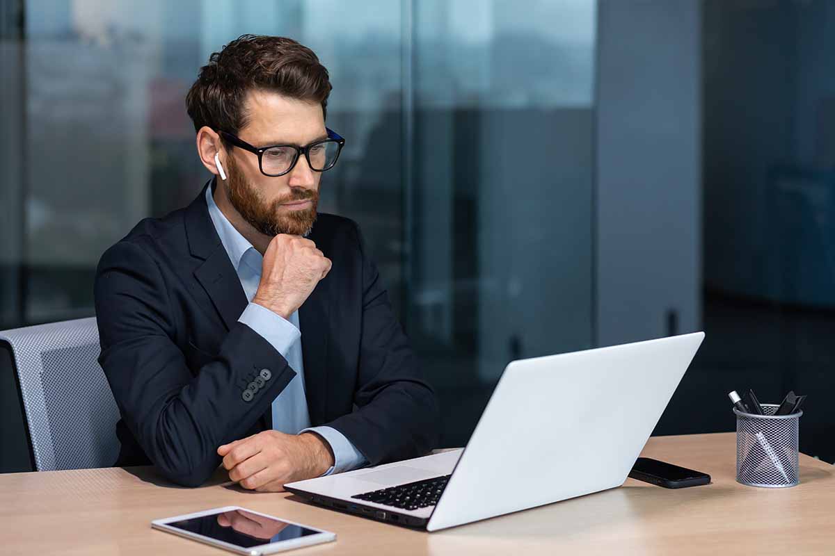 A serious thinking businessman in a navy suit working on a laptop in an office setting.